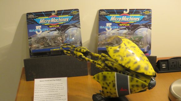 Three toys (two Micro Machines in wrapping in background, one yellow toy in foreground), displayed on a wooden table with explanatory text (included in this essay) next to them.