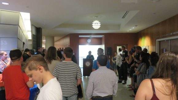 A crowd stands in a hallway. In the background, a woman stands at a podium speaking.
