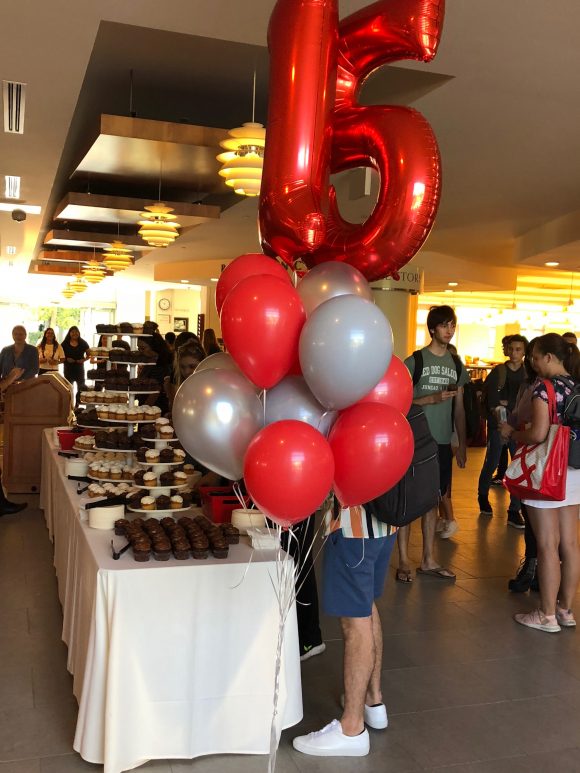 A cluster of red and gray balloons, with two number balloons spelling out "15," stand in front of a table filled with cupcakes.