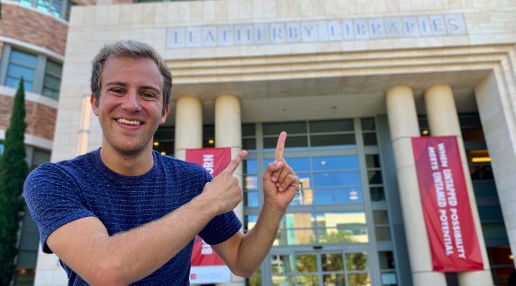 A young man in a blue t-shirt stands outside the front of the Leatherby Libraries, pointing with both his index fingers at the name on the front of the library
