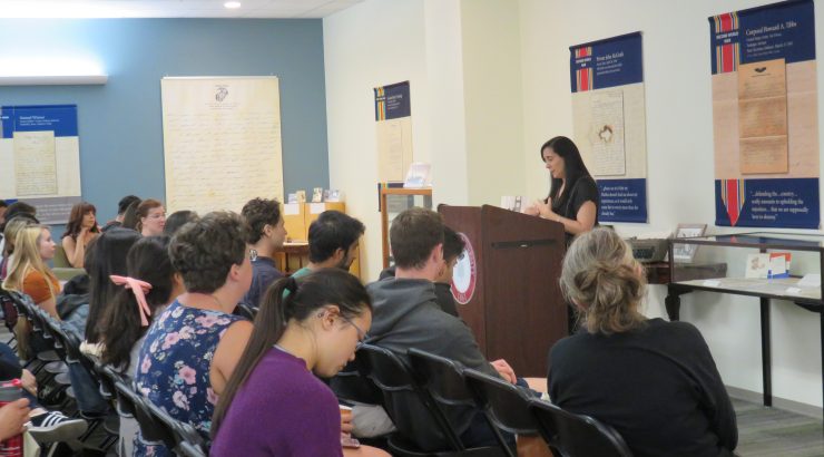 A crowd of people in the foreground sit listening to a woman speaking at a podium in the righthand background of the image