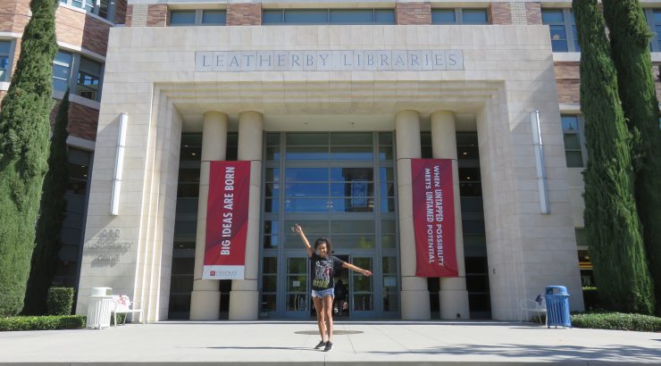 A young woman poses with one arm out and the other up in front of the main entrance to the Leatherby Libraries