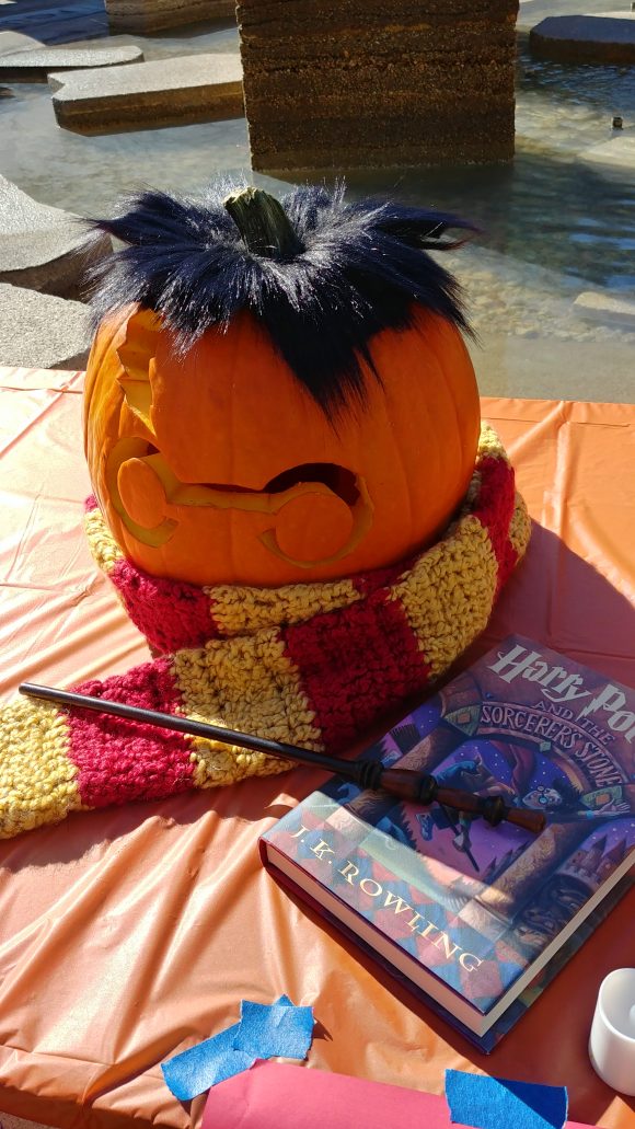 A pumpkin carved to resemble Harry Potter, with the silhouette of the character's glasses and scar, with black fake hair on top, and a red and gold scarf wrapped around the bottom, with a wand and copy of the first Harry Potter book displayed near it.