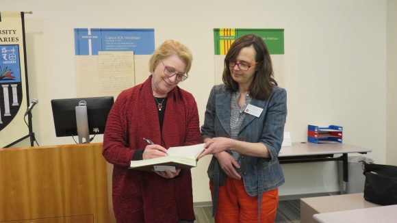 Two women stand next to each other; the woman on the left signs a book while the woman on the right holds the book open and looks on.