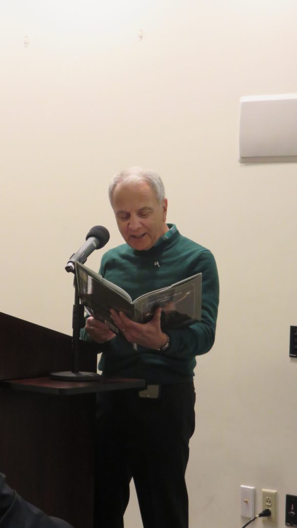 A man stands behind a lectern holding open and reading from a large picture book