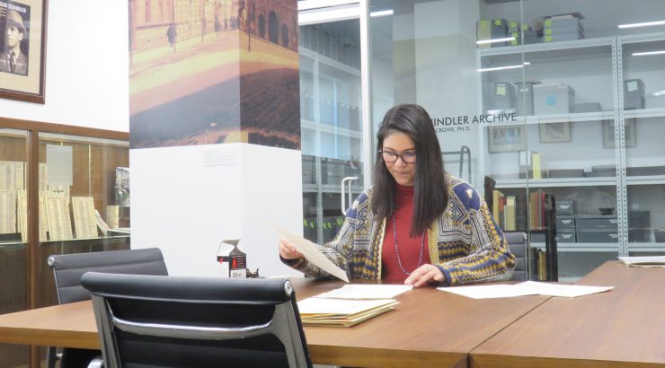 Archivist Tiana Taliep sits reading papers at a table. A glass door behind her is the entrance to the Oskar Schindler Archives.