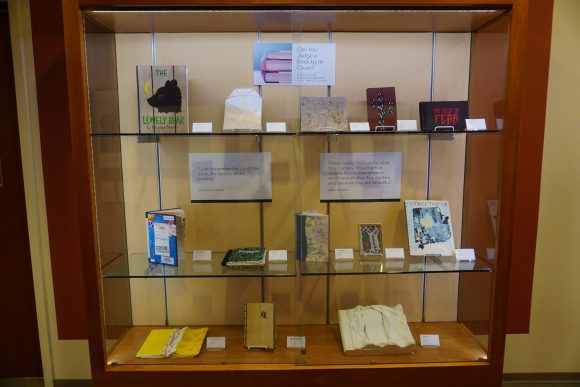 A display case with three glass shelves, each containing a number of artists' books.