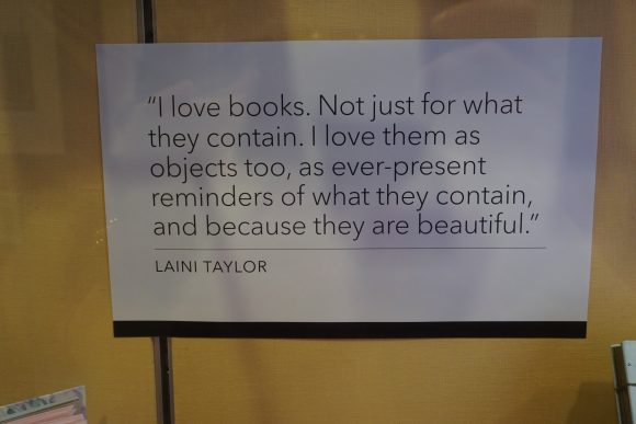 A piece of paper in a display, with text that reads "I love books. Not just for what they contain. I love them as objects, too, as ever-present reminders of what they contain, and because they are beautiful. - Laini Taylor."