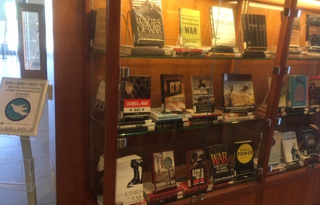 A wood and glass display case containing approximately 20 books, all on the topic of war