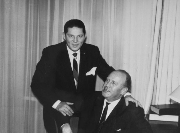 Black and white photograph of two men in suits, one seated and one standing behind him with a hand on the seated man's shoulder