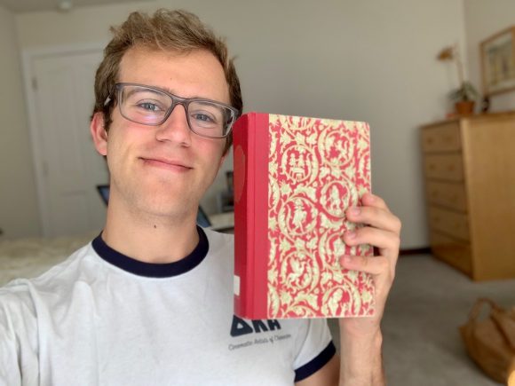 A young blonde man wearing glasses and a white and blue t-shirt holds up a red and yellow patterned hardback book