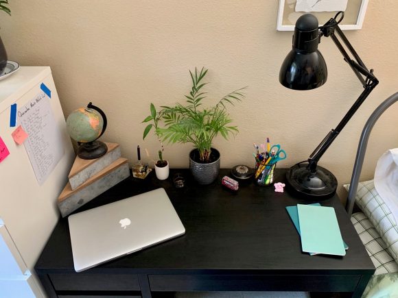 A black desk, with a silver Apple laptop, a mini globe, three small plants, a black lamp, a few desk tools, and two bright blue pieces of paper on it