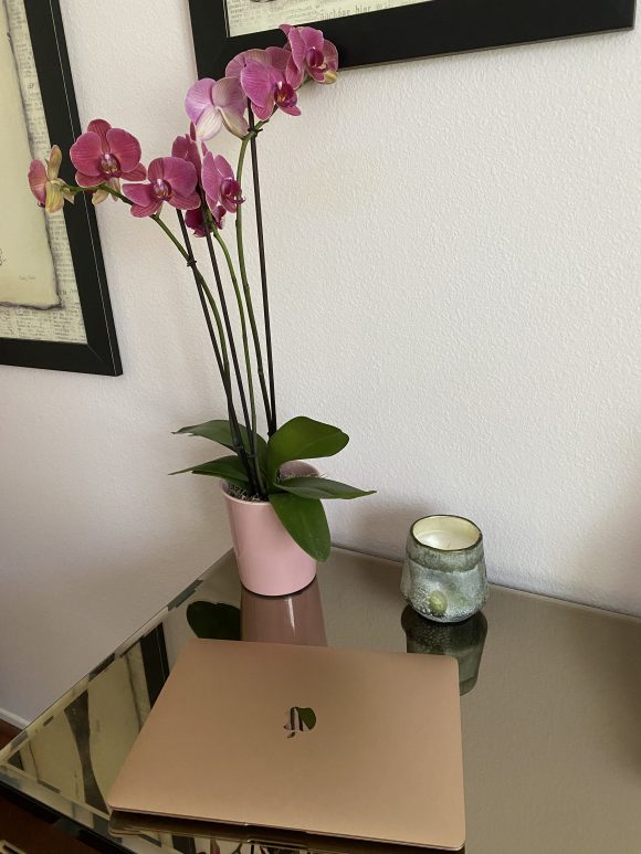A rose gold Apple laptop sitting on a glass-top table, with a candle and a potted pink orchid behind it,