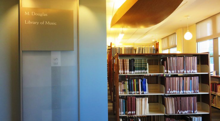 Photograph of the M. Douglas Library of Music in the Leatherby Libraries. A pillar with a sign bearing the name of the library is on the left, and bookshelves filled with books are in the background on the right