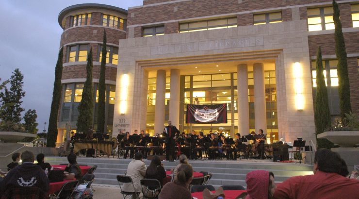 Exterior of the Leatherby Libraries at dusk with an orchestra playing on the stage, a Homecoming banner across the doors, and audience members seated at tables in the Attallah Piazza.