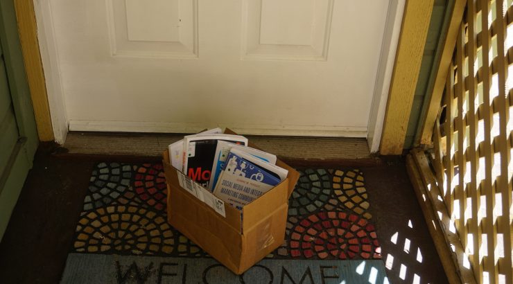 A cardboard box with library books standing up inside it sits on a Welcome mat in front of a door.