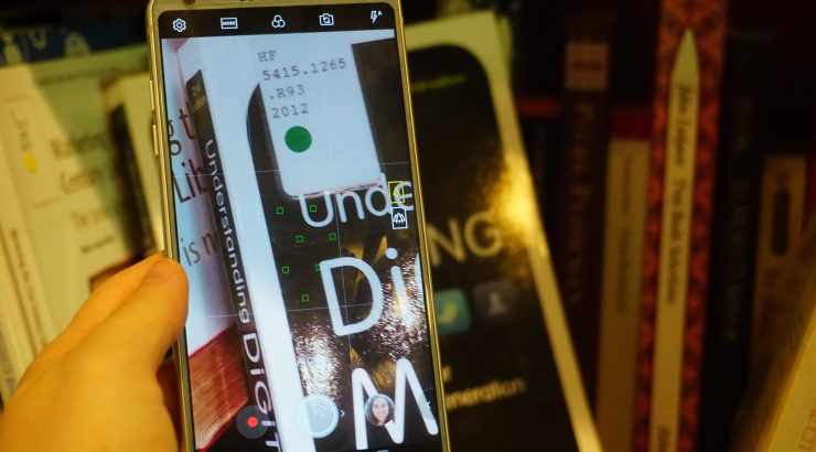 Close-up of a hand holding a cell phone, which is open to the camera feature and zoomed in on the library call number sticker on a book on a bookshelf.
