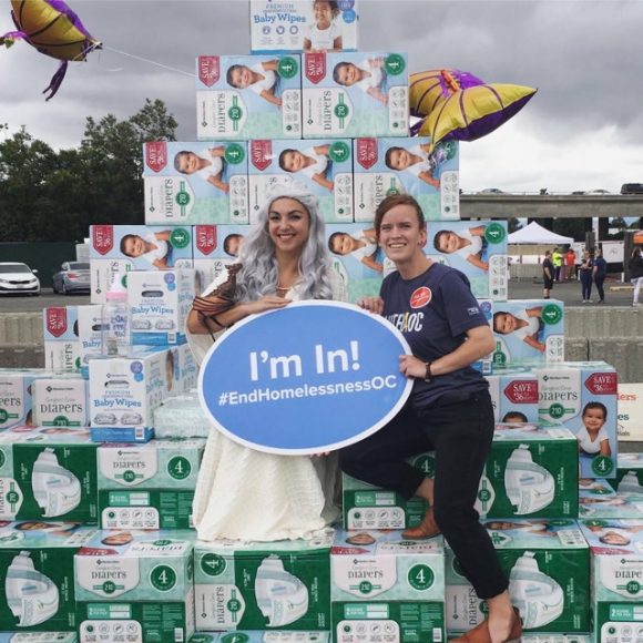 Two people standing in front of diaper pile holding sign saying "Im In! #EndHomelessNessOC"