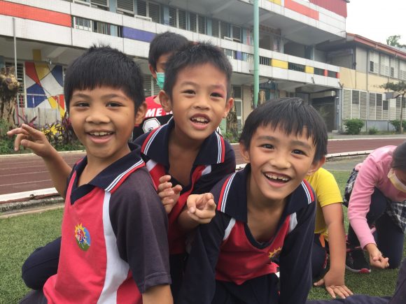 Three young children smiling for the camera in front of their school