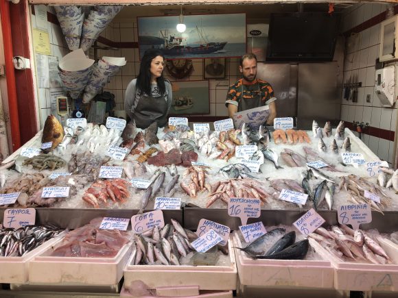 Food stand with variety of fish (red and grey, with skin and filleted, whole fish) over ice with 2 salesmen behind (female on left, male on right).