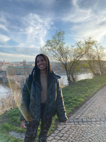 Maia standing on a pathway next to a lake. There is bridge in the background and faded view of the city. The sky is blue with scattered, light clouds and the sun is gleaning on the picture. Maia is smiling and looking to the side towards the lake.