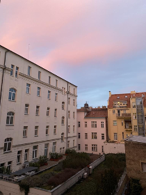 White building on the left with windows on each floor. Yellow building on the top of the photo with similar windows. The picture seems to be taken from the third floor of a building looking out into the street. The sky is cloudy and the clouds have a pink tint to them.