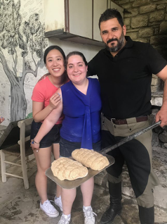 Tiffany smiling and hugging two Greek chefs showing off freshly-made bread.