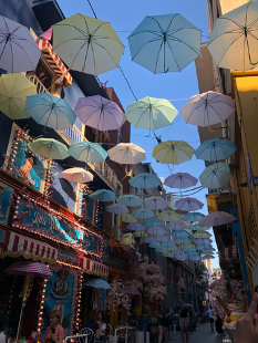 Narrow street with carnival lights and fluorescent colors and lightly colored umbrellas hanging overhead as a decorative ceiling above the street and connected by the roofs of the buildings on each side.