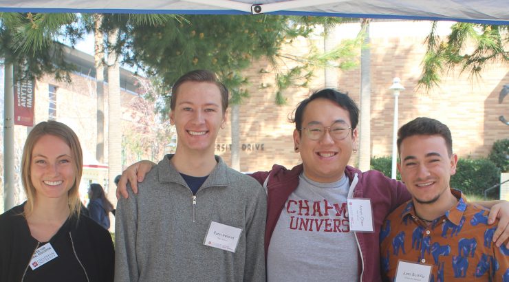 4 student workers standing at a table during study abroad fair. They are hugging each other, smiling, and looking at the camera.