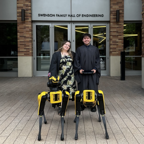 Ethan Tapia (right) with Kalin Richardson (left), dressed in black commencement gowns, posing with the SPOT dogs in front of Swenson Hall